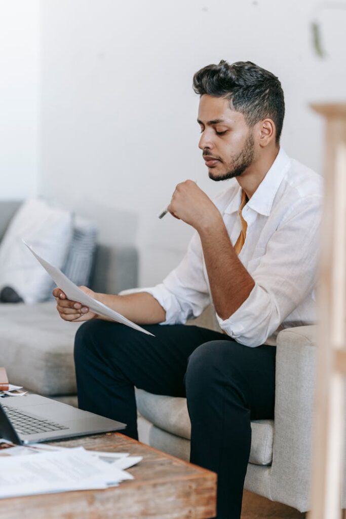 Concentrated young ethnic male wearing casual clothes reading documents and sitting on armchair near table with netbook in living room